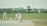 The image captures a group of people engaged in a cricket match. A batsman, wearing a white uniform, is in the act of playing a shot. Behind the batsman, a wicketkeeper and a fielder are positioned, also in white uniforms. The cricket field is green with a boundary line in the background. Beyond the field, there are buildings and a tree, with a banner visible on the right side.