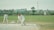 The image captures a group of people engaged in a cricket match. A batsman, wearing a white uniform, is in the act of playing a shot. Behind the batsman, a wicketkeeper and a fielder are positioned, also in white uniforms. The cricket field is green with a boundary line in the background. Beyond the field, there are buildings and a tree, with a banner visible on the right side.