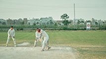 The image captures a group of people engaged in a cricket match. A batsman, wearing a white uniform, is in the act of playing a shot. Behind the batsman, a wicketkeeper and a fielder are positioned, also in white uniforms. The cricket field is green with a boundary line in the background. Beyond the field, there are buildings and a tree, with a banner visible on the right side.