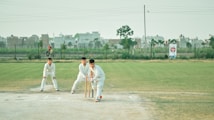 Three individuals dressed in white cricket gear are on a cricket field. One person is in a batting stance with a bat and wickets behind them, while another person is standing nearby in a protective stance. In the background, urban buildings and a person riding a bicycle can be seen. The field has a mix of grass and dirt patches, and the sky is clear with light clouds.