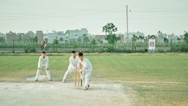 Three individuals dressed in white cricket gear are on a cricket field. One person is in a batting stance with a bat and wickets behind them, while another person is standing nearby in a protective stance. In the background, urban buildings and a person riding a bicycle can be seen. The field has a mix of grass and dirt patches, and the sky is clear with light clouds.