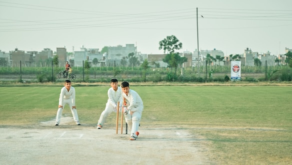 Three individuals dressed in white cricket gear are on a cricket field. One person is in a batting stance with a bat and wickets behind them, while another person is standing nearby in a protective stance. In the background, urban buildings and a person riding a bicycle can be seen. The field has a mix of grass and dirt patches, and the sky is clear with light clouds.