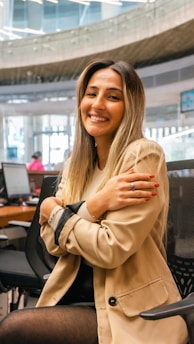 A person with long hair, smiling, is sitting in an office environment. They are wearing a tan blazer and black outfit, with hands crossed over their chest. The background features office furniture and another person working at a desk.