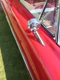 A close-up view of a shiny, vintage red car with chrome detailing, focusing on the side mirror and a portion of the window. The car's glossy surface reflects light, emphasizing its polished finish.