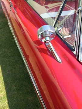 A close-up view of a shiny, vintage red car with chrome detailing, focusing on the side mirror and a portion of the window. The car's glossy surface reflects light, emphasizing its polished finish.