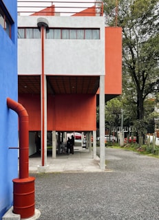 A modern building with a minimalistic architectural style, featuring a vibrant blue and orange color scheme. The structure has large rectangular shapes supported by concrete pillars and includes an external metal stairway. The surroundings are well-kept with trees in the background, and a couple of people are gathered under the building's overhang.
