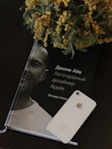 A book featuring a monochrome portrait of a man with Russian text on the cover is placed on a dark surface. Next to it is a white iPhone, and a vibrant floral arrangement with yellow flowers lies partially over the book.