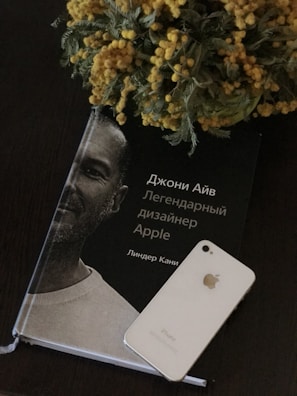 A book featuring a monochrome portrait of a man with Russian text on the cover is placed on a dark surface. Next to it is a white iPhone, and a vibrant floral arrangement with yellow flowers lies partially over the book.