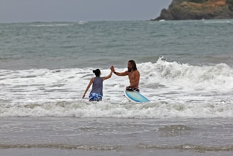 A man and a child are in the ocean, engaging in a high-five. The man is holding a surfboard, and waves are crashing around them. The background features a rocky shoreline under an overcast sky.