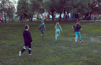 Four children are playing in a grassy park area, surrounded by trees. They appear to be running towards a play area in the background. The park is lush and green, suggesting a lively outdoor setting. People can be seen in the distance near playground equipment.