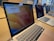 Several open MacBook laptops are displayed on a wooden table in a store setting. The laptops are turned on, showing desktop screens with one displaying a price banner for the MacBook Air. The environment appears to be well-lit with natural light coming through the windows.