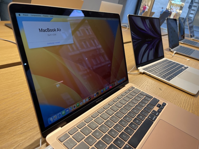 Several open MacBook laptops are displayed on a wooden table in a store setting. The laptops are turned on, showing desktop screens with one displaying a price banner for the MacBook Air. The environment appears to be well-lit with natural light coming through the windows.