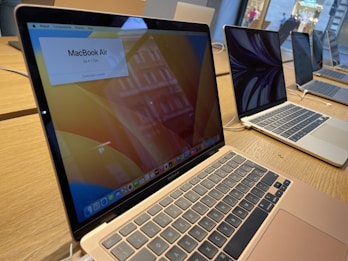 Several open MacBook laptops are displayed on a wooden table in a store setting. The laptops are turned on, showing desktop screens with one displaying a price banner for the MacBook Air. The environment appears to be well-lit with natural light coming through the windows.