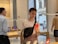 A person holding a red smartphone stands inside a modern store, possibly an electronics or tech store. They are wearing a casual outfit with a crossbody bag. Other people are in the background examining products. Tables display various electronic devices under a sleek, modern interior design with large windows showing an outside street scene.