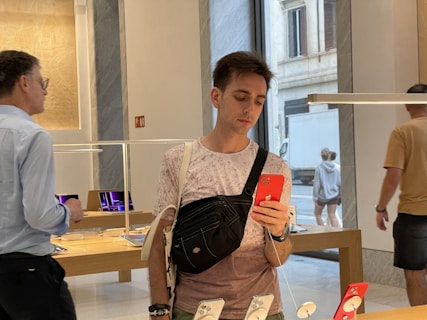 A person holding a red smartphone stands inside a modern store, possibly an electronics or tech store. They are wearing a casual outfit with a crossbody bag. Other people are in the background examining products. Tables display various electronic devices under a sleek, modern interior design with large windows showing an outside street scene.