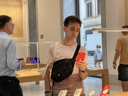 A person holding a red smartphone stands inside a modern store, possibly an electronics or tech store. They are wearing a casual outfit with a crossbody bag. Other people are in the background examining products. Tables display various electronic devices under a sleek, modern interior design with large windows showing an outside street scene.