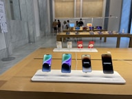 A modern retail store interior featuring a display of mobile phones on wooden tables. The foreground has a row of four smartphones on display, each with its own charging station and information label. In the background, people are browsing other electronic devices, possibly tablets, on another table.