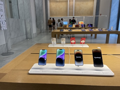 A modern retail store interior featuring a display of mobile phones on wooden tables. The foreground has a row of four smartphones on display, each with its own charging station and information label. In the background, people are browsing other electronic devices, possibly tablets, on another table.