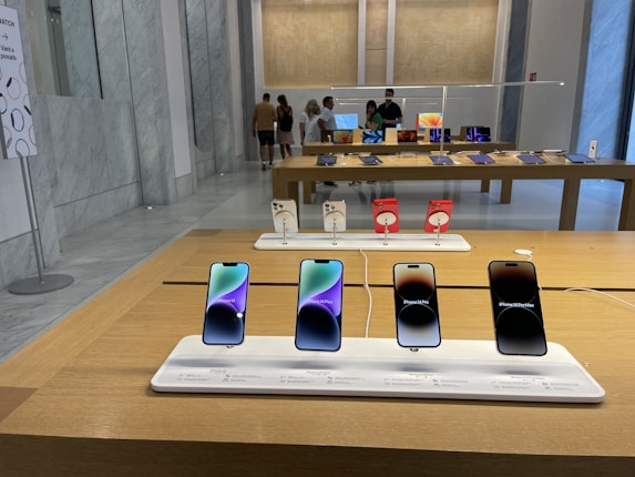 A modern retail store interior featuring a display of mobile phones on wooden tables. The foreground has a row of four smartphones on display, each with its own charging station and information label. In the background, people are browsing other electronic devices, possibly tablets, on another table.