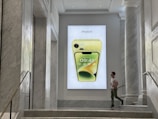 A man is walking past a large advertisement featuring a yellow iPhone 14 displayed on a wall. The setting is an elegant interior with marble walls and staircases, giving a modern, luxurious feel.