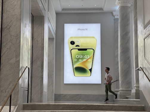 A man is walking past a large advertisement featuring a yellow iPhone 14 displayed on a wall. The setting is an elegant interior with marble walls and staircases, giving a modern, luxurious feel.