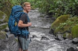 A person smiles while standing next to a fast-flowing stream in a lush, green forest environment. They are carrying a large blue backpack equipped with climbing gear and wearing casual outdoor attire.