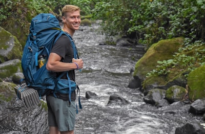A person smiles while standing next to a fast-flowing stream in a lush, green forest environment. They are carrying a large blue backpack equipped with climbing gear and wearing casual outdoor attire.