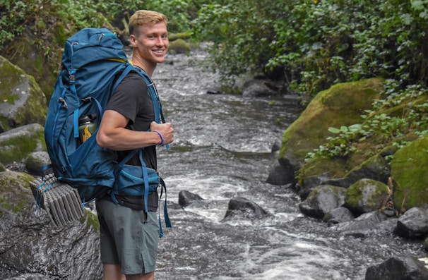 A person smiles while standing next to a fast-flowing stream in a lush, green forest environment. They are carrying a large blue backpack equipped with climbing gear and wearing casual outdoor attire.