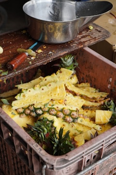A collection of freshly peeled pineapple skins and tops are inside a large plastic crate. Next to it, a wooden table holds a metal bowl and a knife with a wooden handle, suggesting preparation of fruit.