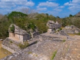 Ancient ruins with several stone structures overgrown with vegetation. The stones are weathered, and the scene is set amidst a lush, green landscape with trees and a bright blue sky filled with clouds, capturing a sense of history and nature intertwined.