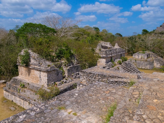 Ancient ruins with several stone structures overgrown with vegetation. The stones are weathered, and the scene is set amidst a lush, green landscape with trees and a bright blue sky filled with clouds, capturing a sense of history and nature intertwined.