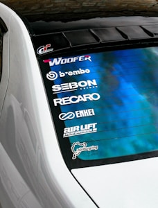The rear window of a white car features several automotive brand decals including Woofer, Brembo, Seibon Carbon, Recaro, Enkei, and Air Lift Performance. The background of the window has a blue gradient design. Additionally, there is a Nürburgring track outline sticker.
