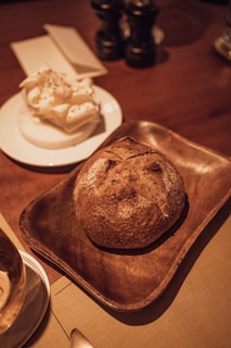 A rustic loaf of bread is placed on a brown wooden tray, accompanied by a plate holding a portion of whipped butter. The setting includes a wooden table with two pepper grinders and a napkin holder in the background. The warm lighting adds a cozy atmosphere to the scene.