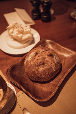 A rustic loaf of bread is placed on a brown wooden tray, accompanied by a plate holding a portion of whipped butter. The setting includes a wooden table with two pepper grinders and a napkin holder in the background. The warm lighting adds a cozy atmosphere to the scene.