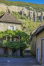 A picturesque stone house covered in lush green vines sits on a narrow street, with a rugged cliff face and dense forest in the background under a clear blue sky.