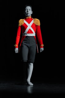 A person wearing a bright red military-style costume with gold fringe shoulder epaulettes and white diagonal straps. The background is dark, highlighting the figure dramatically as they stand with one leg in a dance pose.