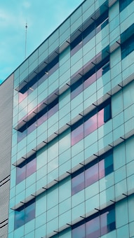 The glass facade of a modern building reflects the sky, featuring a grid of rectangular windows with some horizontal external shades. The structure displays a pattern of alternating tinted panels that create a geometric design.