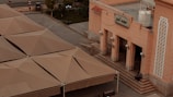 A large building with an ornate facade, featuring intricate patterns and a sign in Arabic script. Broad, beige-colored structures resembling tents or shade roofs cover an adjacent area. A person is seated on steps leading to the building entrance. Street lamps and a water tank are visible nearby, with a few trees and a fence in the background.