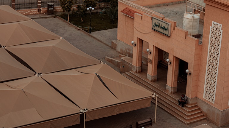 A large building with an ornate facade, featuring intricate patterns and a sign in Arabic script. Broad, beige-colored structures resembling tents or shade roofs cover an adjacent area. A person is seated on steps leading to the building entrance. Street lamps and a water tank are visible nearby, with a few trees and a fence in the background.