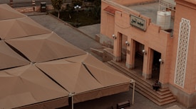 A large building with an ornate facade, featuring intricate patterns and a sign in Arabic script. Broad, beige-colored structures resembling tents or shade roofs cover an adjacent area. A person is seated on steps leading to the building entrance. Street lamps and a water tank are visible nearby, with a few trees and a fence in the background.