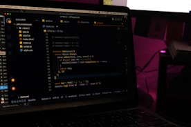 A laptop screen displays a code editor with several JavaScript files open in the sidebar, including app2.js, app.js, index.html, index.js, script.js, and style.css. The editor shows a JavaScript function with syntax highlighting, and the environment appears dimly lit with a dark mode interface. Several folders and files are visible within the editor's explorer pane.