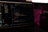 A laptop screen displays a code editor with several JavaScript files open in the sidebar, including app2.js, app.js, index.html, index.js, script.js, and style.css. The editor shows a JavaScript function with syntax highlighting, and the environment appears dimly lit with a dark mode interface. Several folders and files are visible within the editor's explorer pane.