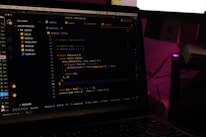 A laptop screen displays a code editor with several JavaScript files open in the sidebar, including app2.js, app.js, index.html, index.js, script.js, and style.css. The editor shows a JavaScript function with syntax highlighting, and the environment appears dimly lit with a dark mode interface. Several folders and files are visible within the editor's explorer pane.