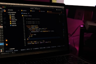 A laptop screen displays a code editor with several JavaScript files open in the sidebar, including app2.js, app.js, index.html, index.js, script.js, and style.css. The editor shows a JavaScript function with syntax highlighting, and the environment appears dimly lit with a dark mode interface. Several folders and files are visible within the editor's explorer pane.