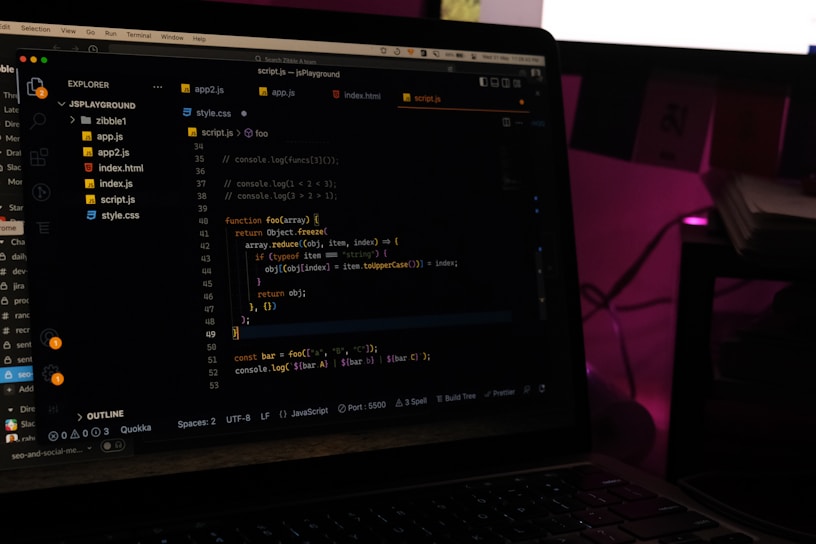 A laptop screen displays a code editor with several JavaScript files open in the sidebar, including app2.js, app.js, index.html, index.js, script.js, and style.css. The editor shows a JavaScript function with syntax highlighting, and the environment appears dimly lit with a dark mode interface. Several folders and files are visible within the editor's explorer pane.