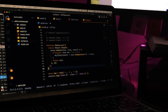 A laptop screen displays a code editor with several JavaScript files open in the sidebar, including app2.js, app.js, index.html, index.js, script.js, and style.css. The editor shows a JavaScript function with syntax highlighting, and the environment appears dimly lit with a dark mode interface. Several folders and files are visible within the editor's explorer pane.
