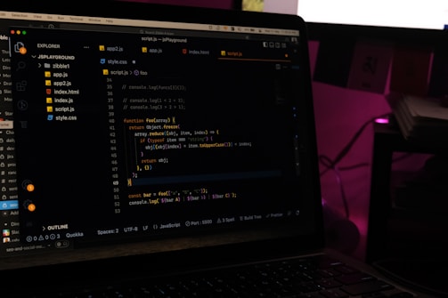 A laptop screen displays a code editor with several JavaScript files open in the sidebar, including app2.js, app.js, index.html, index.js, script.js, and style.css. The editor shows a JavaScript function with syntax highlighting, and the environment appears dimly lit with a dark mode interface. Several folders and files are visible within the editor's explorer pane.