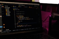 A laptop screen displays a code editor with several JavaScript files open in the sidebar, including app2.js, app.js, index.html, index.js, script.js, and style.css. The editor shows a JavaScript function with syntax highlighting, and the environment appears dimly lit with a dark mode interface. Several folders and files are visible within the editor's explorer pane.