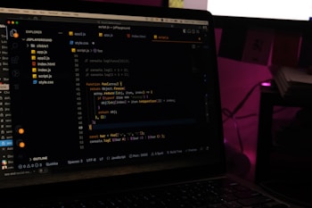 A laptop screen displays a code editor with several JavaScript files open in the sidebar, including app2.js, app.js, index.html, index.js, script.js, and style.css. The editor shows a JavaScript function with syntax highlighting, and the environment appears dimly lit with a dark mode interface. Several folders and files are visible within the editor's explorer pane.