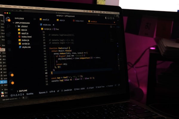 A laptop screen displays a code editor with several JavaScript files open in the sidebar, including app2.js, app.js, index.html, index.js, script.js, and style.css. The editor shows a JavaScript function with syntax highlighting, and the environment appears dimly lit with a dark mode interface. Several folders and files are visible within the editor's explorer pane.